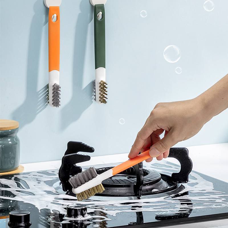 A hand is using an orange kitchen gas stove cleaning brush with plastic scraper on a black gas stove top.