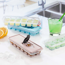 Load image into Gallery viewer, An ice cube tray with a lid in a light cashmere color, displayed on a kitchen countertop with ice cubes inside it. A glass with a green liquid and a lemon slice are also visible in the background.
