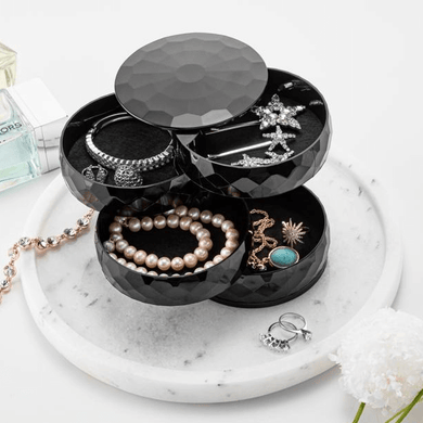 A black, diamond-shaped, multi-tiered jewelry and accessories box with various items such as bracelets, rings, and a necklace displayed on a marble surface.