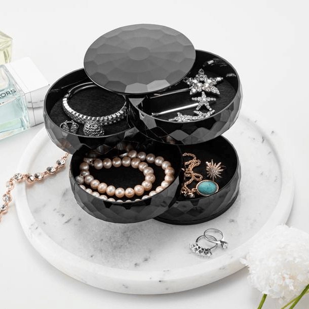 A black, diamond-shaped, multi-tiered jewelry and accessories box with various items such as bracelets, rings, and a necklace displayed on a marble surface.