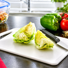 Load image into Gallery viewer, Two halves of a green lettuce wedge on a white cutting board with a knife, set on a dark kitchen countertop with whole vegetables and a window in the background.
