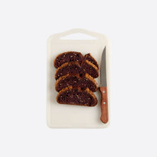Load image into Gallery viewer, Sliced dark brown bread on a white cutting board with a knife on a white background
