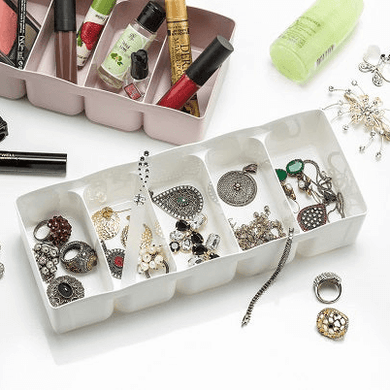 A white plastic storage organizer with 5 compartments, shown partially filled with various small items such as jewelry and cosmetic products, placed on a surface with more cosmetic products and jewelry scattered around it.